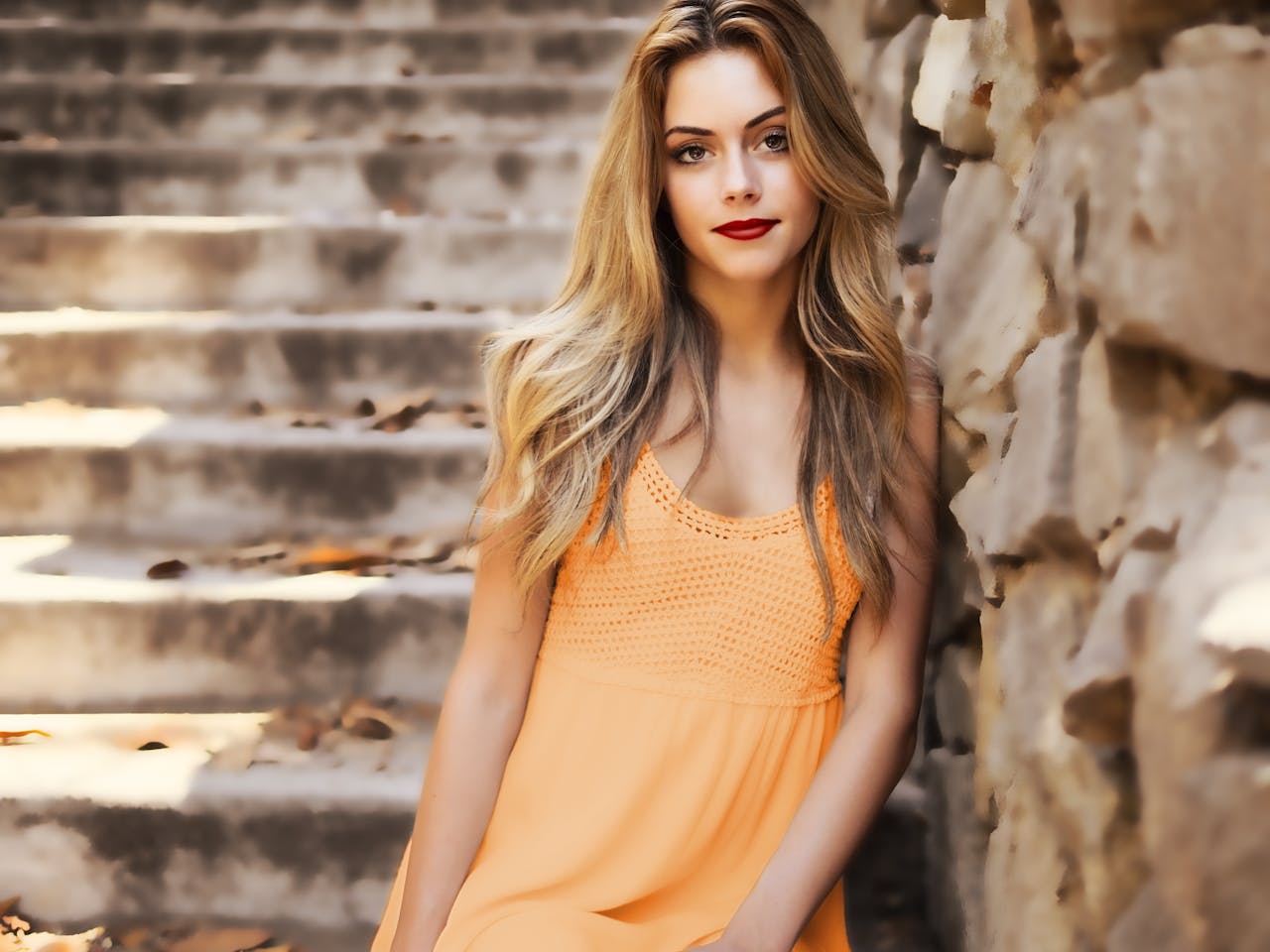 A beautiful woman with long blonde hair and red lips posing by a stone wall outdoors.