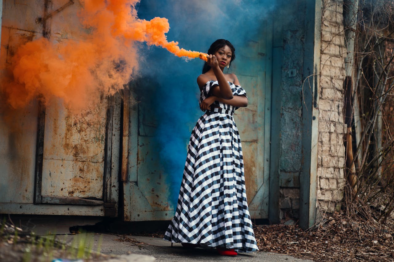 A stylish woman in a checkered dress poses with vibrant smoke outdoors against a rustic backdrop.